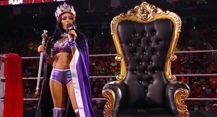 Adorning a sceptre and wearing a cape and crown, Zelina Vega cuts a promo in front of a throne.