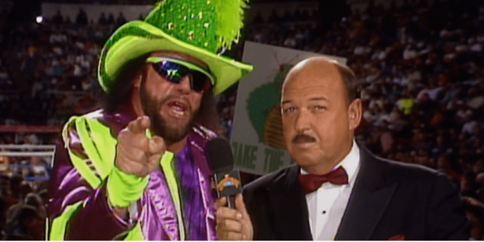 Randy Savage talks into the mic, pointing at the camera as he is decked in a sequined, feathery hat and bright green and purple stripes.
