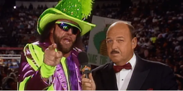 Randy Savage talks into the mic, pointing at the camera as he is decked in a sequined, feathery hat and bright green and purple stripes.