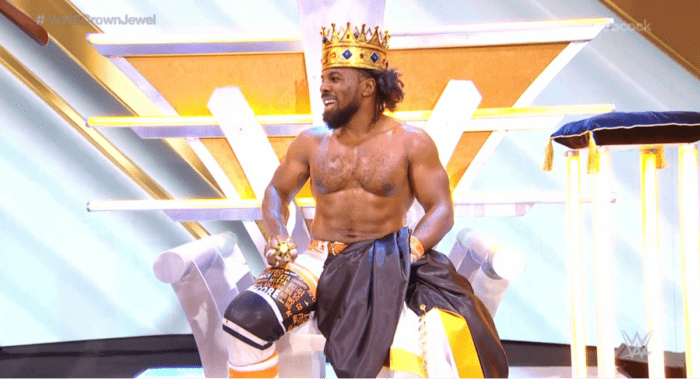 Xavier Woods sits on the stage, crown on head.