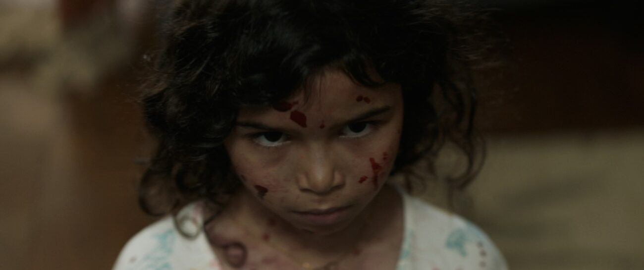 A child stands with their head tilted, and a grimace on their blood spattered face.