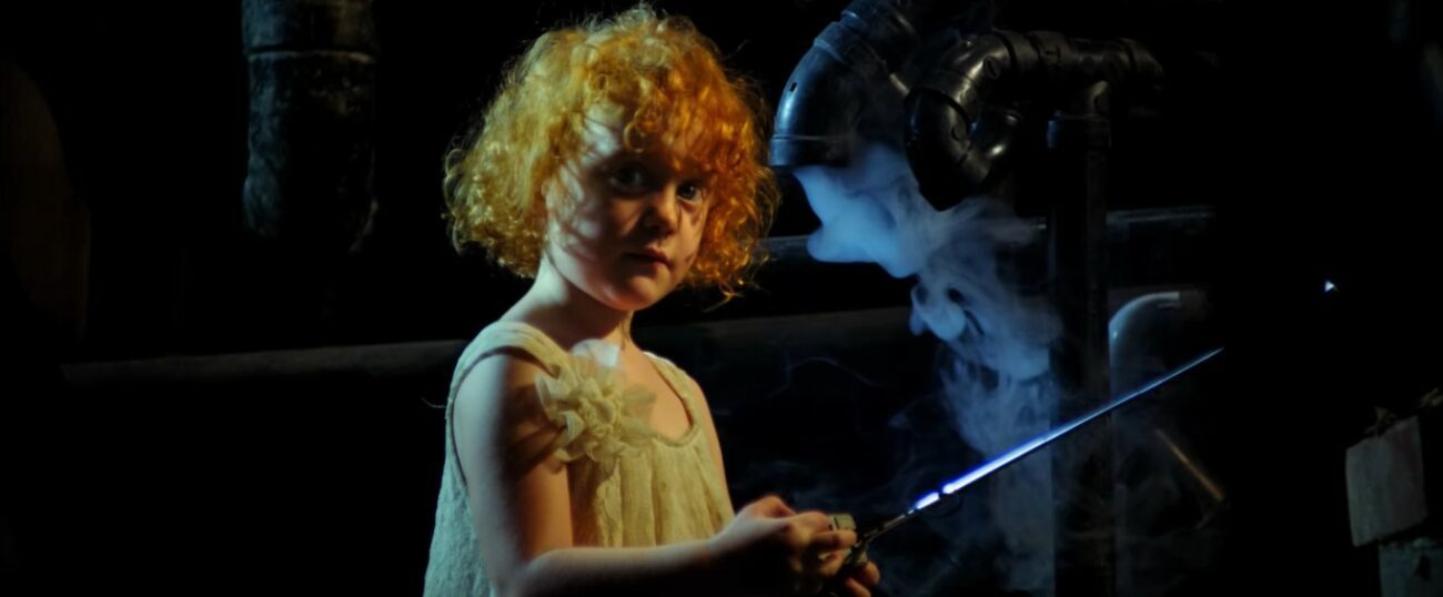 A young girl looks towards the camera, the shadows of her hair fall over her face. A pipe in the background emits an ominous white fog.