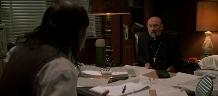 A priest and a professor sit across from each other at a desk. The table is covered with scattered papers.