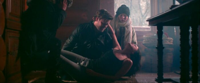 A woman is placed on the floor of a cabin by a handsome man while a man and woman crowd around