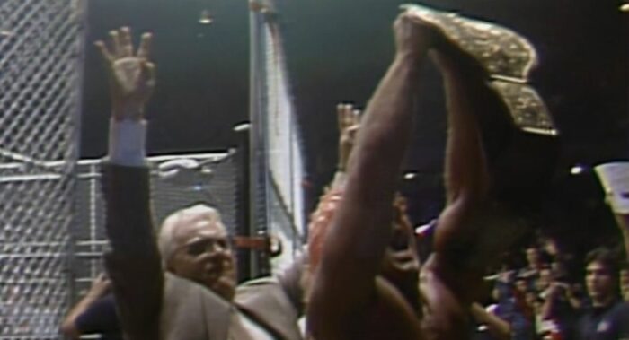 A bloody Ric Flair holds aloft the Big Gold Belt outside the steel cage as J.J. Dillon hold four fingers aloft.