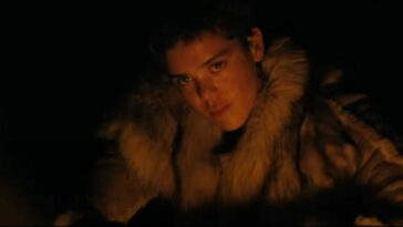 a woman in a fur coat looks toward the camera as the light from a fire reflects off of her face in "Out of Darkness'