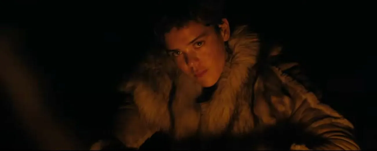 a woman in a fur coat looks toward the camera as the light from a fire reflects off of her face in "Out of Darkness'