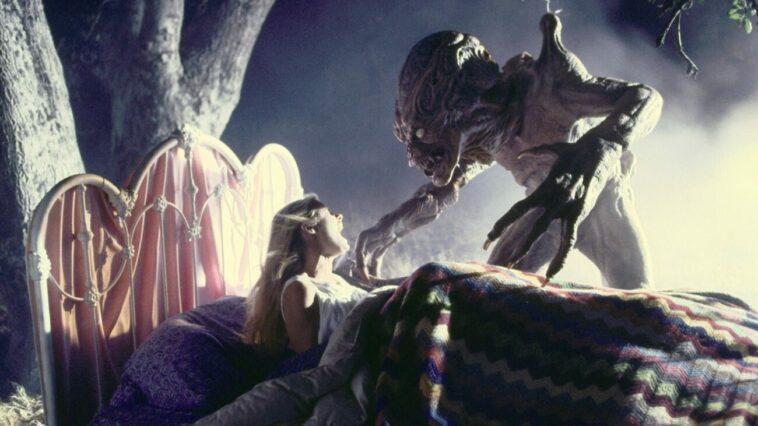 Pumpkinhead stands over a woman in a bed, who is in the middle of the woods