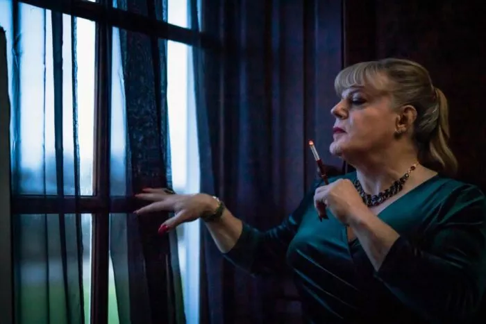 Nina looks through the curtains of a large window while smoking a cigarette.