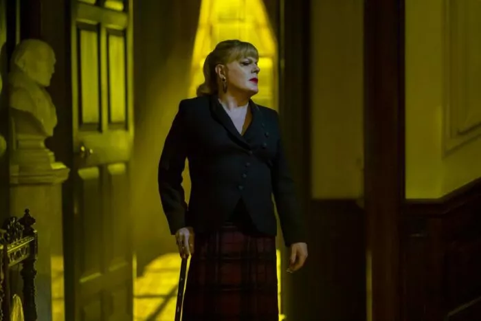 Nina stands in the yellow glow of the outside light from the window, looking to her left while holding a cane in Doctor Jekyll