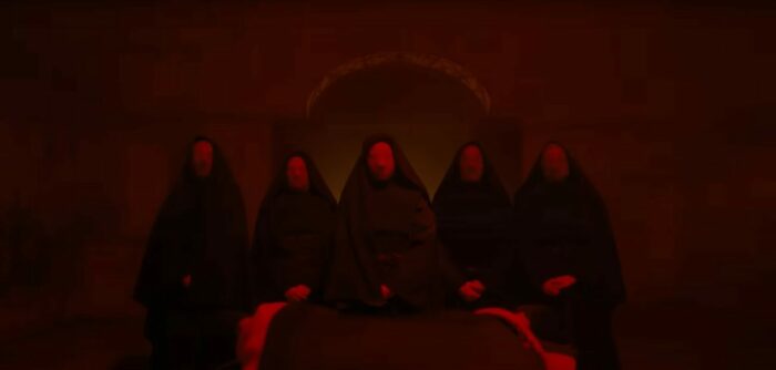 A row of five red veiled nuns stand in a red room over Cecilia in Immaculate