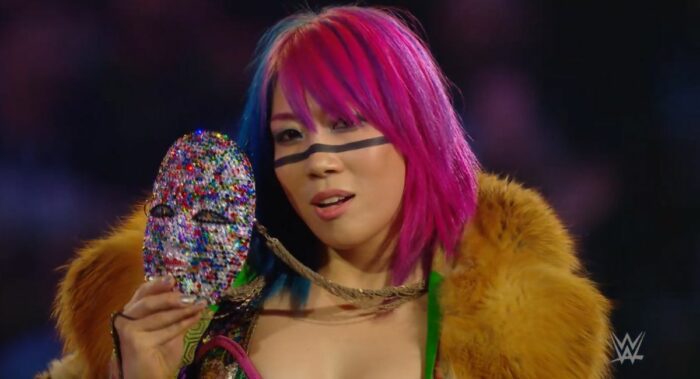Asuka holds up her jewelled mask and gazes into the camera.