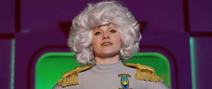 A woman with a bouffant in a space uniform against a purple and green background