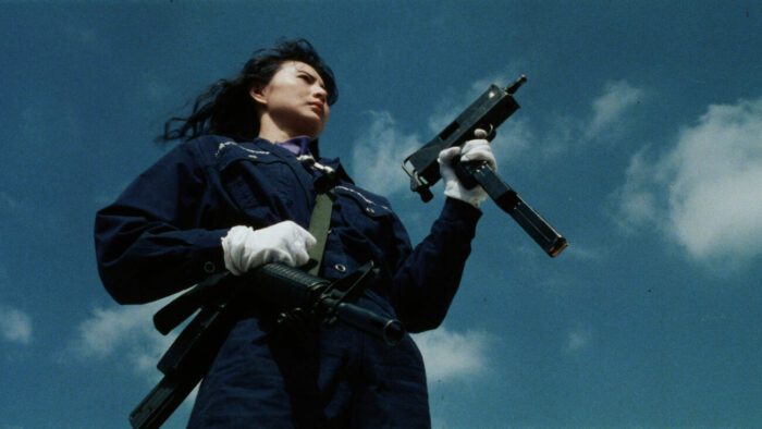 A woman in coveralls with a canvas bag strapped across her body holds an uzi against a blue sky