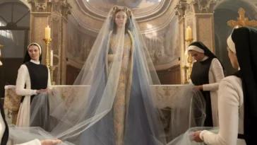 Nuns surround a veiled woman standing in a gold gown with a blue cloth over her and a golden crown of flowers stands before an altar in a cathedral