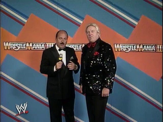 Mean Gene and Bobby Heenan stand in front of the garish WrestleMania III backstage backdrop