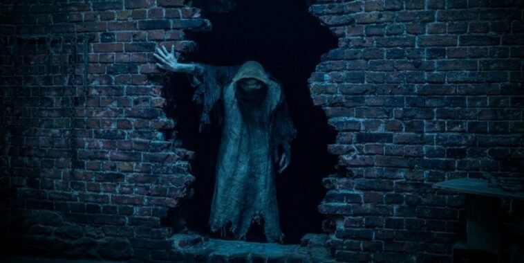 A hooded figure emerges from a hole in a brick wall in Baghead.