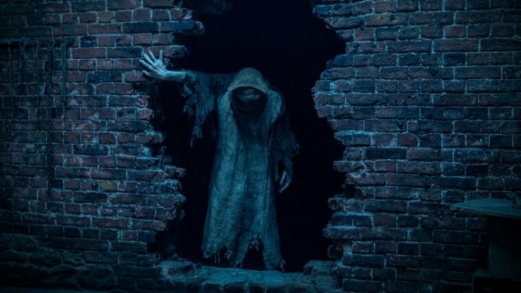 A hooded figure emerges from a hole in a brick wall in Baghead.