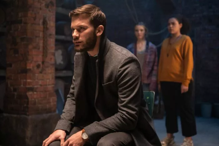A man in a peacoat crouches down in a basement while two women look over his shoulder.