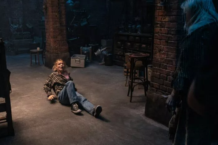 A woman on her back on a cement floor looks forward in terror as a figure approaches