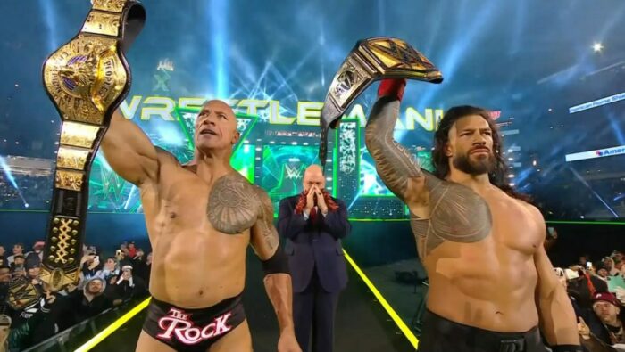 The Rock holding his People's Champion belt next to Roman Reigns holding his Undisputed WWE Universal Championship, with Paul Heyman in the middle