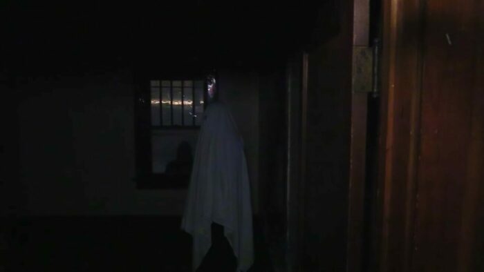 A man standing beneath a sheet pretending to be a ghost in a dark empty room while a shadowy figure is seen through the window