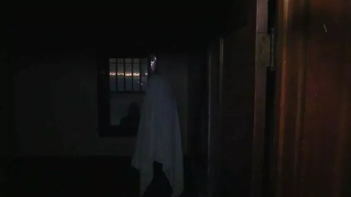 A man standing beneath a sheet pretending to be a ghost in a dark empty room while a shadowy figure is seen through the window