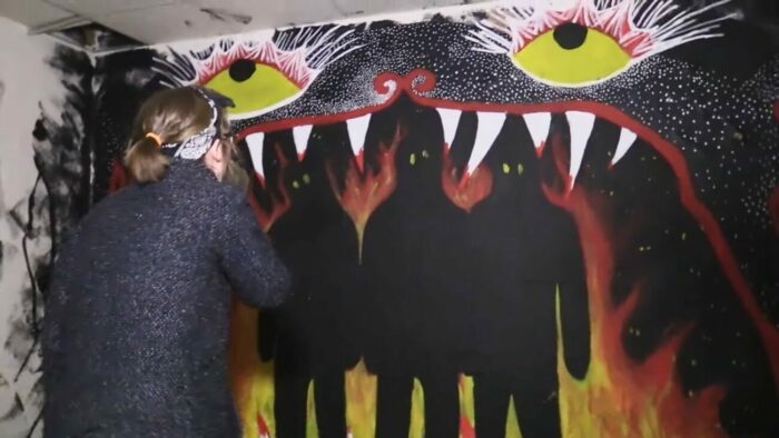 a woman covers her mouth at the painting of three shadow figures with yellow eyes standing among flames inside a creatures large fanged mouth.