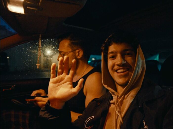 two men sit in a car, one is waving at the camera and smiling