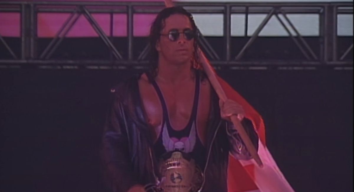 Bret Hart, in John Lennon sunglasses, has the WWF belt around his waist and flag over shoulder.