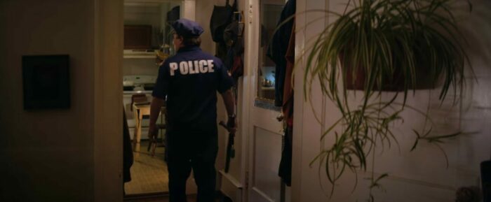 A man in a police uniform holding a police baton stands in the doorway of a home in Sweet Relief.
