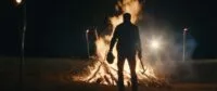 A man standing in front of a fire, holding a head and an axe