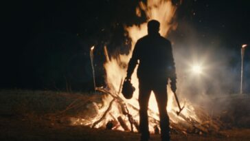 A man standing in front of a fire, holding a head and an axe