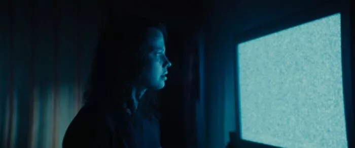 The glow of a static-filled television light the face of a girl in a dark room.