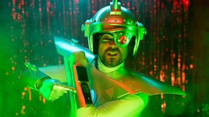 A space man in a metal helmet covering one eye points to his electronic armband in The Wheel of Heaven