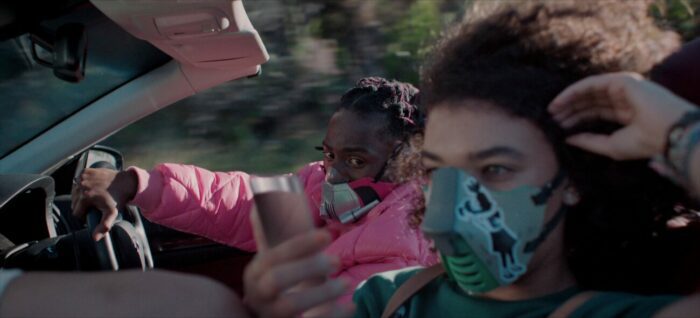 A man in a pink windbreaker drives a convertable with a mask on next to a woman holding her phone, also wearing a mask.