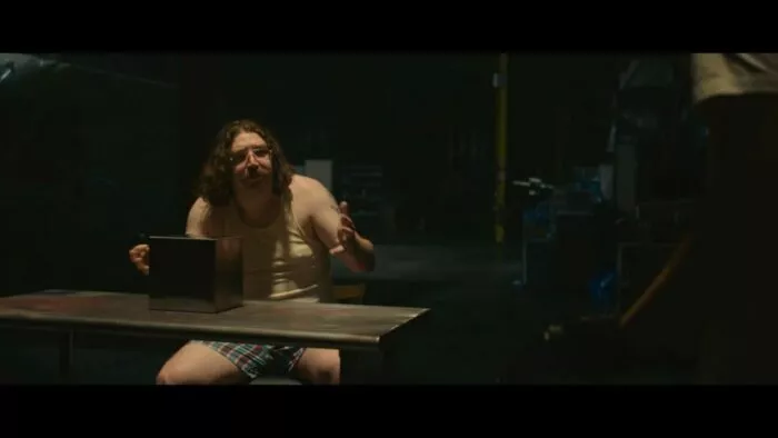 A long haired man in glasses speaks animatedly with his hands while sitting at a cube device on a table.