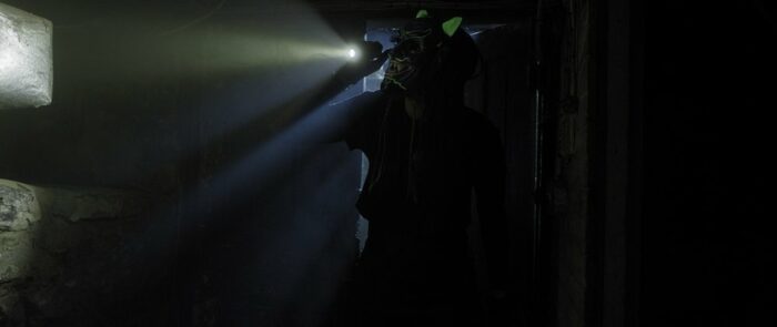 A masked person with a flashlight enters a dark room