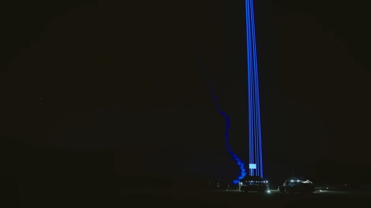 Multiple lasers aim toward the sky with the trail of rocket smoke illuminated behind it