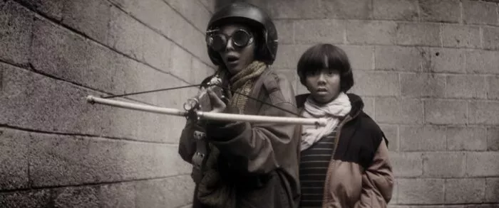 A small boy hides behind a slighlty older boy wearing goggles and a helmet and holding a crossbow in PARVULOS
