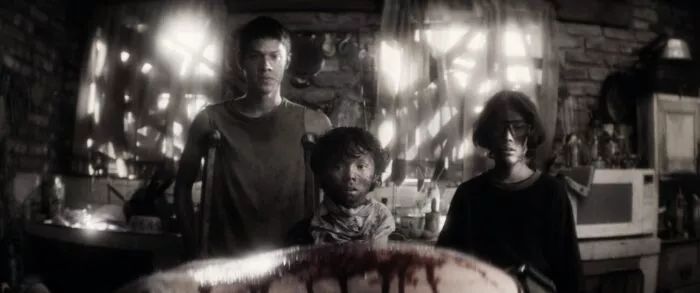 Three dirty looking boys stand on the opposite side of a kitchen table where a bloodied bowl rests in front of them, while light streaks through the boarded up windows behind them.