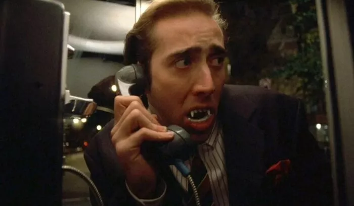 A suited man (Nic Cage) wearing fake vampire fangs looks worried whilst on the phone in a phone booth in Vampire's Kiss (1988)
