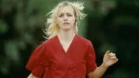A woman in scrubs with blond hair and bleeding from the ear looks upset while running toward the camers in STRANGE DARLING