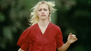 A woman in scrubs with blond hair and bleeding from the ear looks upset while running toward the camers in STRANGE DARLING