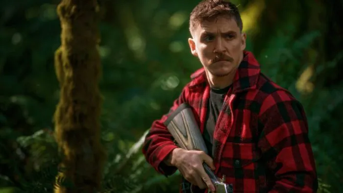 A man with a mustache in a flannel shirt holding a rifle in the woods
