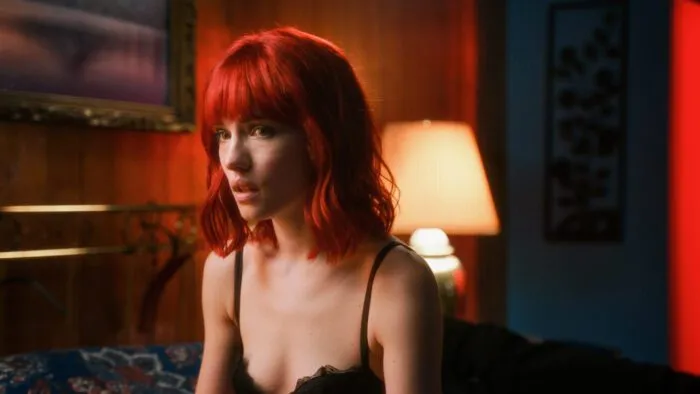 A woman with cherry red hair sits in her bra on the corner of a motel room bed in STRANGE DARLING