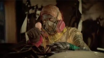 A person with a dirty, soot-covered face in prodective gear and a ventilator face mask stares at the doll head they're holding in ANIMALIA PARADOXA