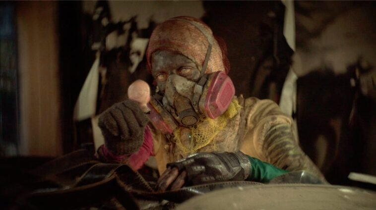 A person with a dirty, soot-covered face in prodective gear and a ventilator face mask stares at the doll head they're holding in ANIMALIA PARADOXA