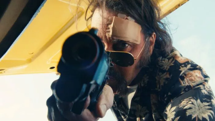 A man with a bandage on his head aims a gun barrel straight into the camera