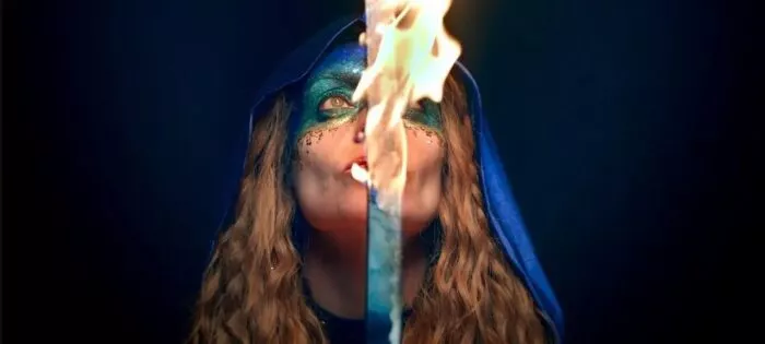 A woman in a blue hoodie and sparkling blue facepaint over her eyes holds a sword on fire